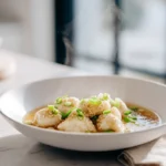 Photorealistic bowl of dumpling soup with steaming broth, dumplings, and green onions on a marble counter in a modern kitchen.